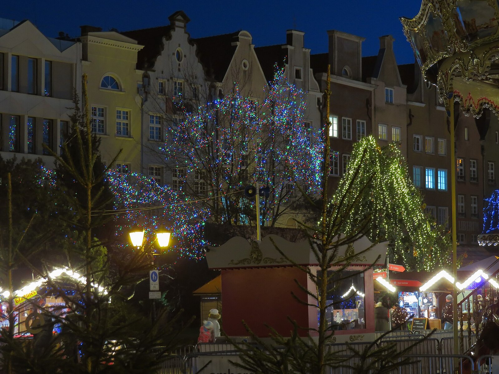 Stalls of the Gdańsk Christmas Market in the evening, lit Christmas trees and the facade of the Great Armoury